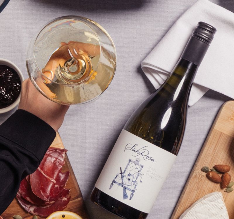 Glass of the 2021 Subrosa grampians viognier, held by a hand. Bottle of the white wine is laying next to the glass, with cheese, nuts and charcuterie nearby.