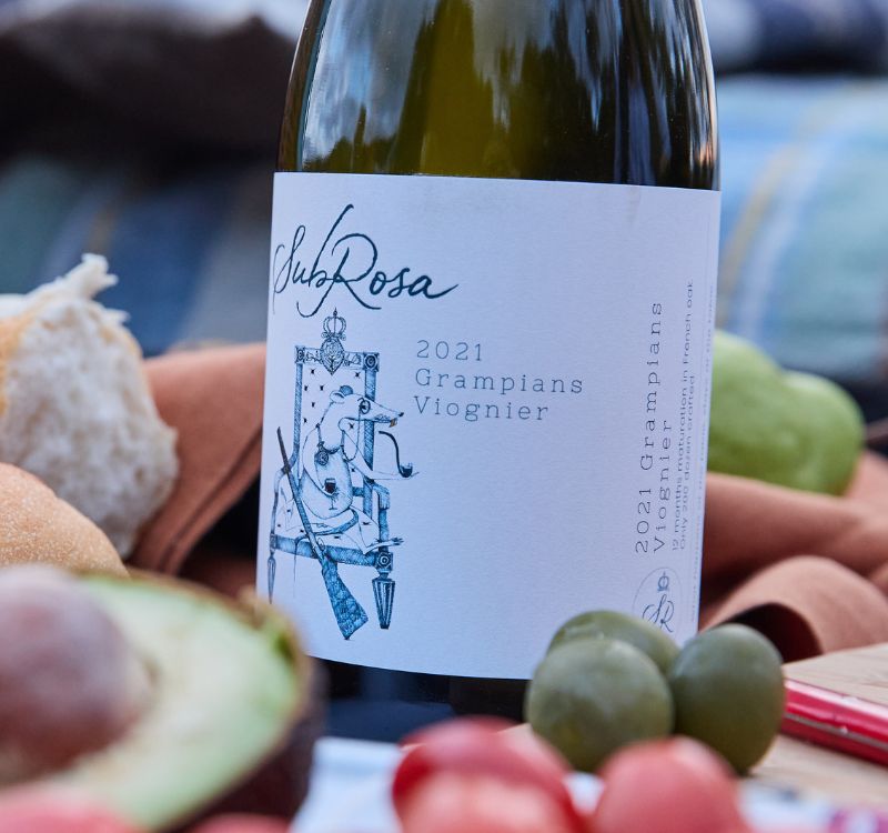Bottle of SubRosa viognier surrounded by food including avocado, tomatoes and olives.